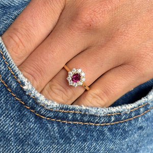 A close-up of a hand wearing a yellow gold ring with a central round pink ruby surrounded by a halo of diamonds, set against blue denim fabric.