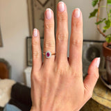 Hand wearing a Vintage 1940s yellow gold ring with a pink ruby at the centre and  a diamond halo, blurred indoor background