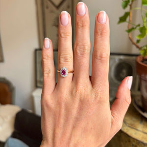 Hand wearing a Vintage 1940s yellow gold ring with a pink ruby at the centre and  a diamond halo, blurred indoor background