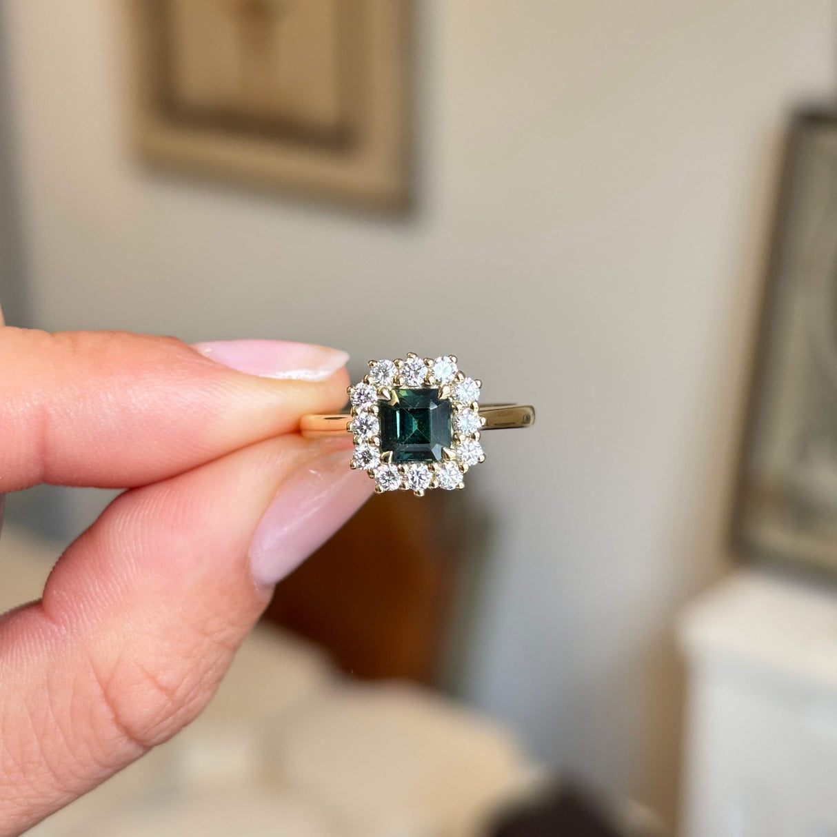 A hand holds an Antique-inspired, teal sapphire and diamond cluster engagement ring set against a softly blurred indoor background.