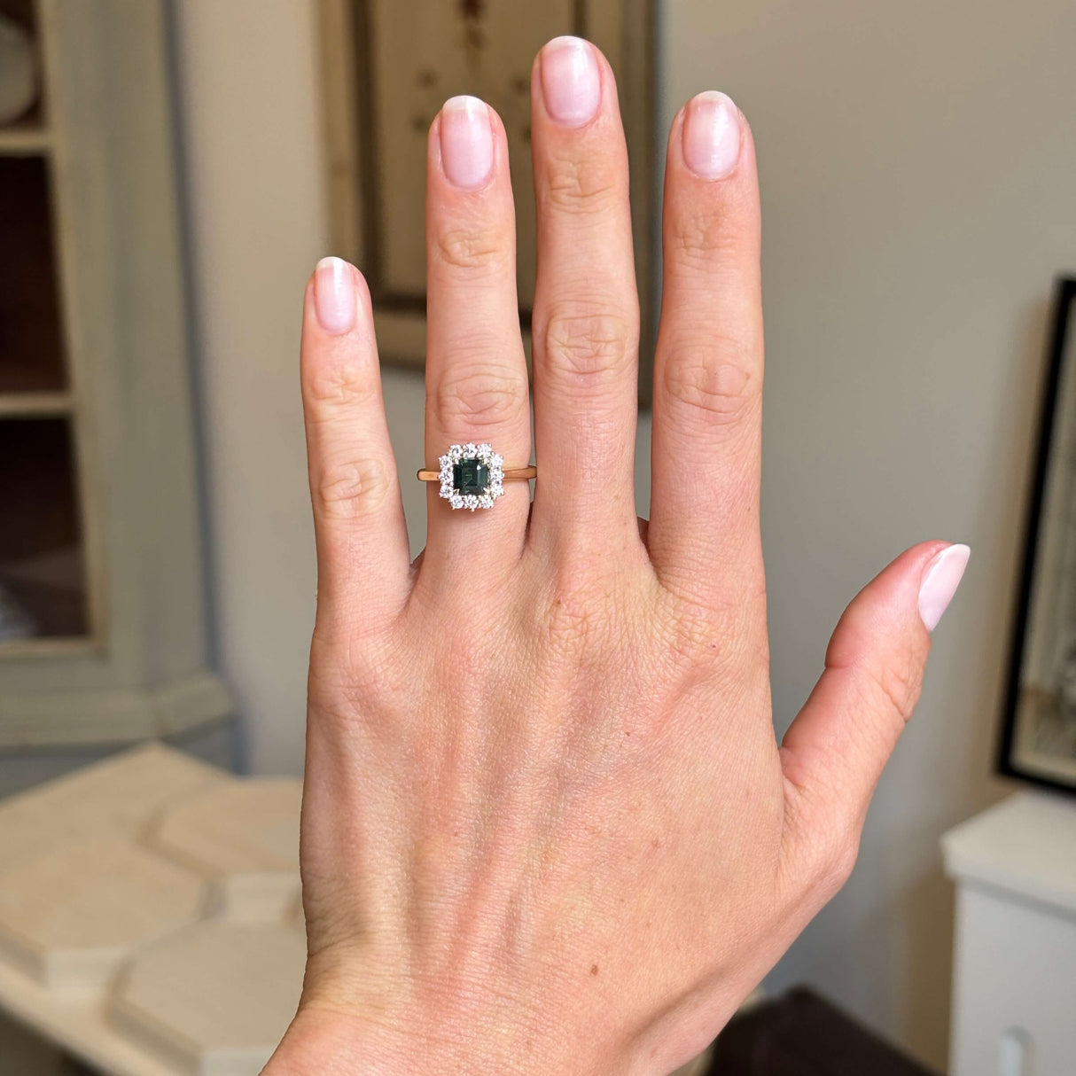 A close-up of a hand wearing an antique-inspired engagement ring with a square-cut green sapphire stone surrounded by a halo of diamonds in a gold setting, with a plain gold band.