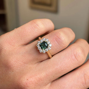 A close-up of a hand wearing an antique-inspired ring with a green rectangular centre sapphire surrounded by a halo of diamonds on a yellow gold band.