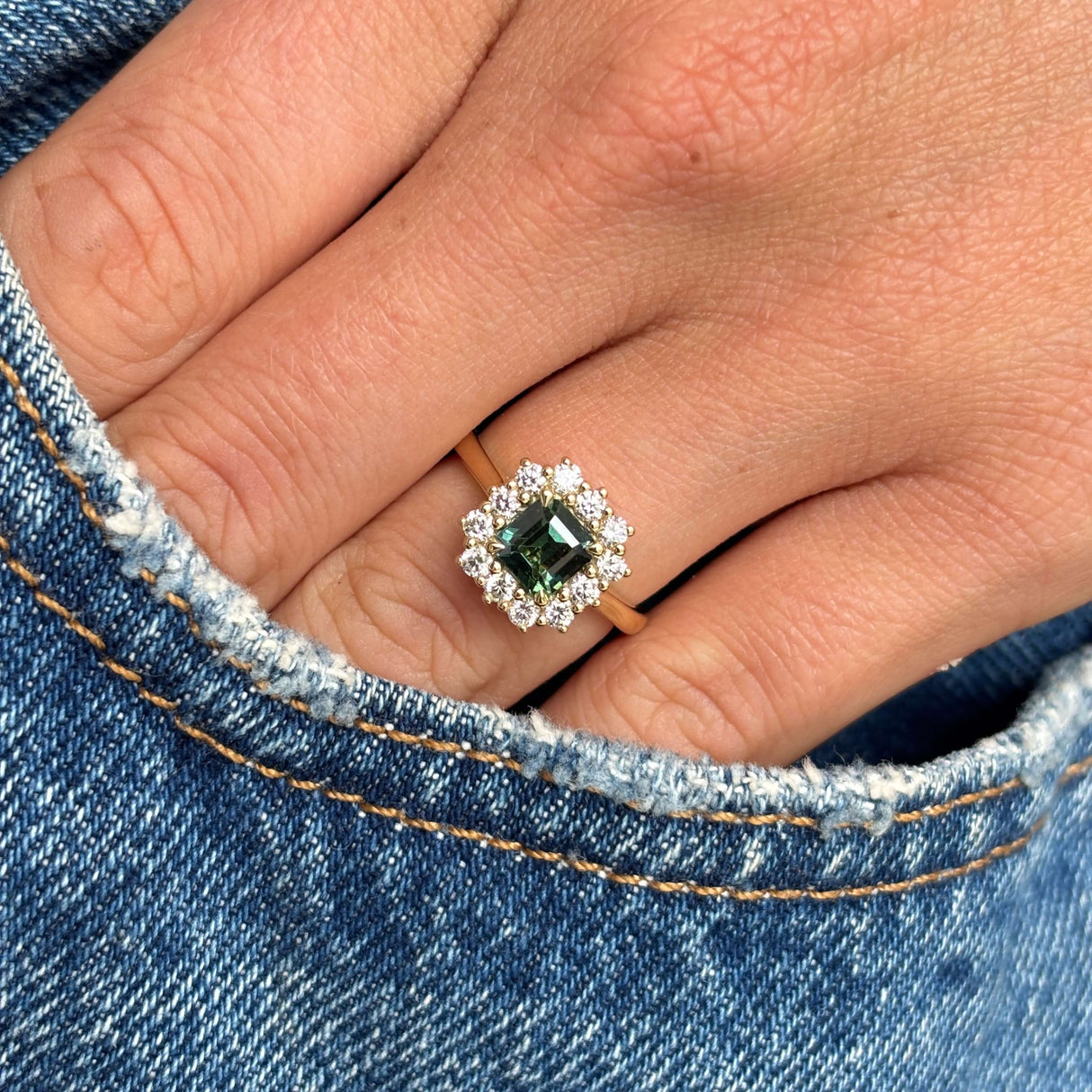 A close-up of a hand wearing an antique-inspired engagement ring with a square green gemstone surrounded by a halo of diamonds on a gold band, set against blue denim fabric.