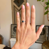 A Vintage-inspired, Green sapphire and diamond cluster trefoil engagement ring, shown on a person's ring finger. The hand is posed against a softly blurred indoor background featuring framed art and a plant.