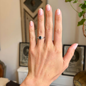 A Vintage-inspired, Green sapphire and diamond cluster trefoil engagement ring, shown on a person's ring finger. The hand is posed against a softly blurred indoor background featuring framed art and a plant.