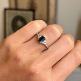 A platinum Vintage-inspired engagement ring with a rectangular green emerald-cut sapphire flanked by diamonds, shown worn on a person's finger.