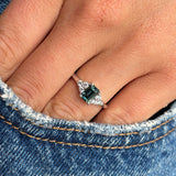 A close-up of a hand wearing a platinum engagement ring with a central emerald-cut green sapphire with diamonds to either side, shown against blue denim fabric.