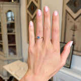 A close-up of a hand wearing an antique inspired blue sapphire & diamond three-stone engagement ring set against a softly blurred indoor background.