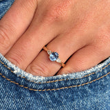 A close-up of a hand wearing an antique-inspired gold engagement ring with a cornflower blue sapphire and two diamonds resting near the pocket of blue denim jeans.