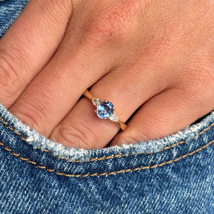 A close-up of a hand wearing an antique-inspired gold engagement ring with a cornflower blue sapphire and two diamonds resting near the pocket of blue denim jeans.
