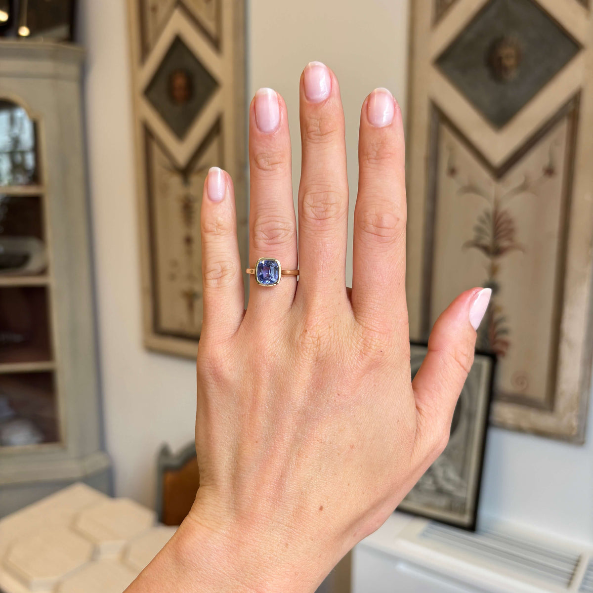 A close-up of a hand wearing an Antique-inspired, violet sapphire solitaire engagement ring in yellow gold, set against an indoor background with framed wall art.