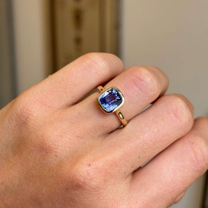 An Antique-inspired, solitaire engagement ring set with a purple cushion-cut sapphire with a yellow gold bezel setting and band, worn on a person's finger, shown in a close-up shot.