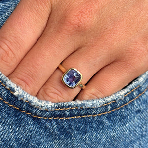 A close-up of a hand wearing a yellow gold bezel set purple sapphire solitaire ring, resting on blue denim jeans.