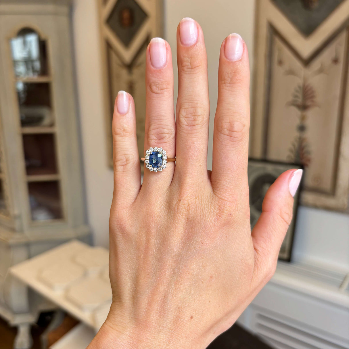 A close-up of a hand wearing an antique-inspired ring with a blue cushion-cut central sapphire, surrounded by a halo of diamonds on a yellow gold band, set against an indoor home background.