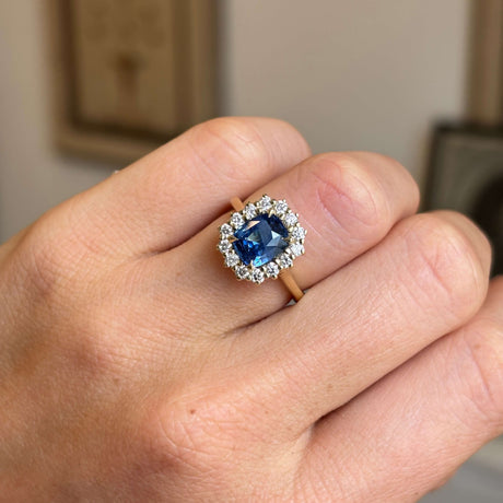 A close-up of a hand wearing an antique-inspired yellow gold engagement ring with a cushion-cut blue sapphire and a halo of diamonds.