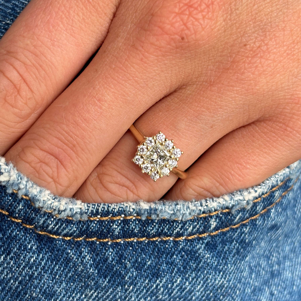 An Antique-inspired, princess-cut diamond cluster engagement ring in yellow gold worn on a hand with a denim background. 