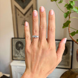 A close-up of a person's hand wearing an Antique, Edwardian gold three-stone diamond ring, with a softly blurred indoor background featuring framed art and a plant.