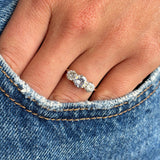 A close-up of a hand wearing an Edwardian gold  three-stone ring set with three old-cut diamonds,  resting on the edge of blue denim jeans.