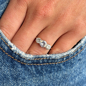 A close-up of a hand wearing an Edwardian gold  three-stone ring set with three old-cut diamonds,  resting on the edge of blue denim jeans.