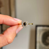 A close-up of a hand holding an antique inspired yellow gold ring with a central dark brown square-cut diamond surrounded by a halo of small diamonds, set against a softly blurred indoor background.