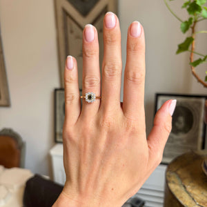 A hand wearing an Antique-inspired, Brown diamond cluster engagement ring in yellow gold, on the wedding finger, set against an interiors background.