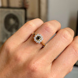 A close-up of a hand wearing an antique-inspired, diamond cluster ring with a brown diamond central stone and a gold band, set against a softly blurred indoor background.