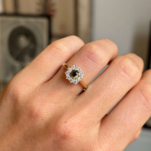 A close-up of a hand wearing an antique-inspired, diamond cluster ring with a brown diamond central stone and a gold band, set against a softly blurred indoor background.