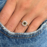 A close-up of a hand wearing an antique-inspired brown green diamond cluster ring with a gold band, resting against blue denim fabric.