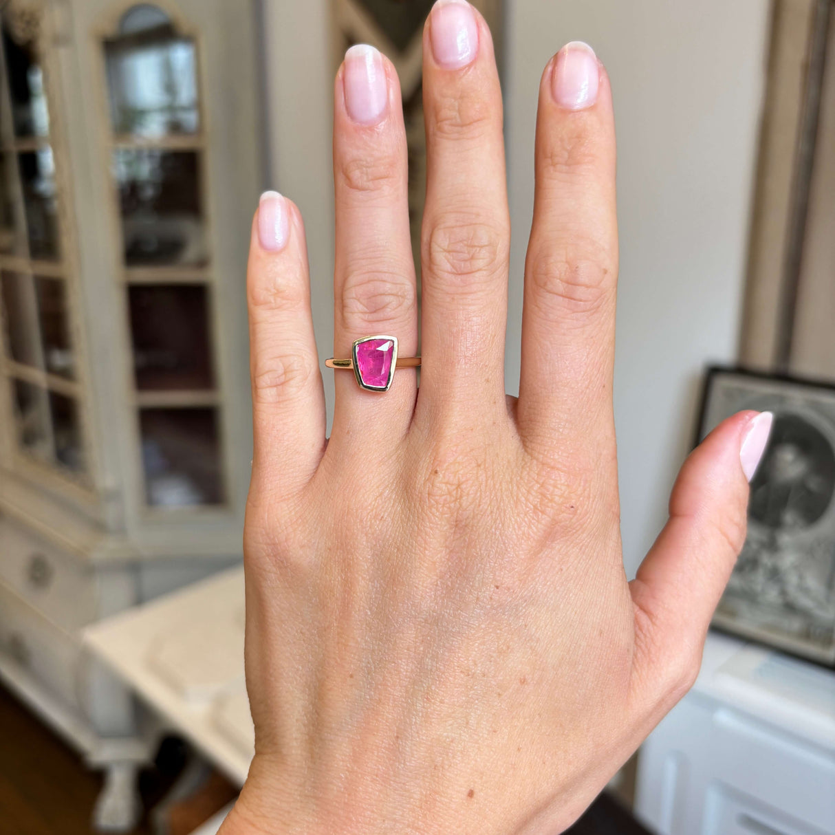 Hand wearing a gold Vintage-inspired, pink ruby solitaire engagement ring against a blurred indoor background
