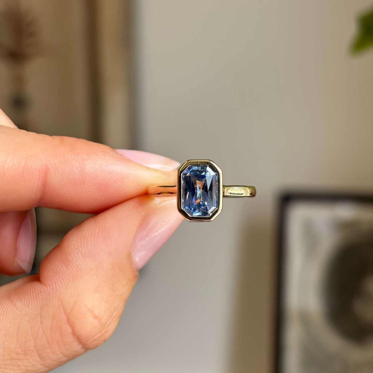 An antique-inspired yellow gold solitaire engagement ring set with a blue sapphire held between fingers against a softly blurred indoor background.