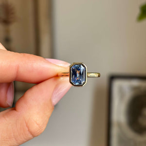 An antique-inspired yellow gold solitaire engagement ring set with a blue sapphire held between fingers against a softly blurred indoor background.