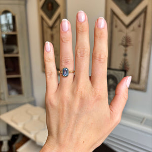An antique-inspired solitaire engagement ring worn on a person's ring finger, displaying the rectangular-cut blue sapphire, shown against a softly blurred indoor background.