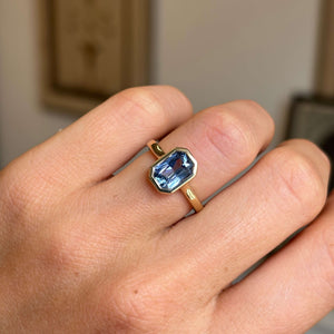An antique-inspired, cornflower blue sapphire solitaire engagement ring in yellow gold, worn on a person's finger.