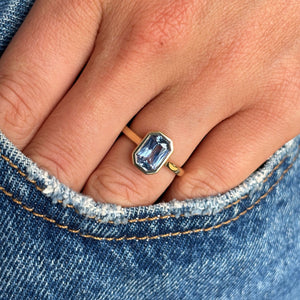 A close-up of a hand wearing a yellow gold blue sapphire engagement ring, resting against blue denim fabric.