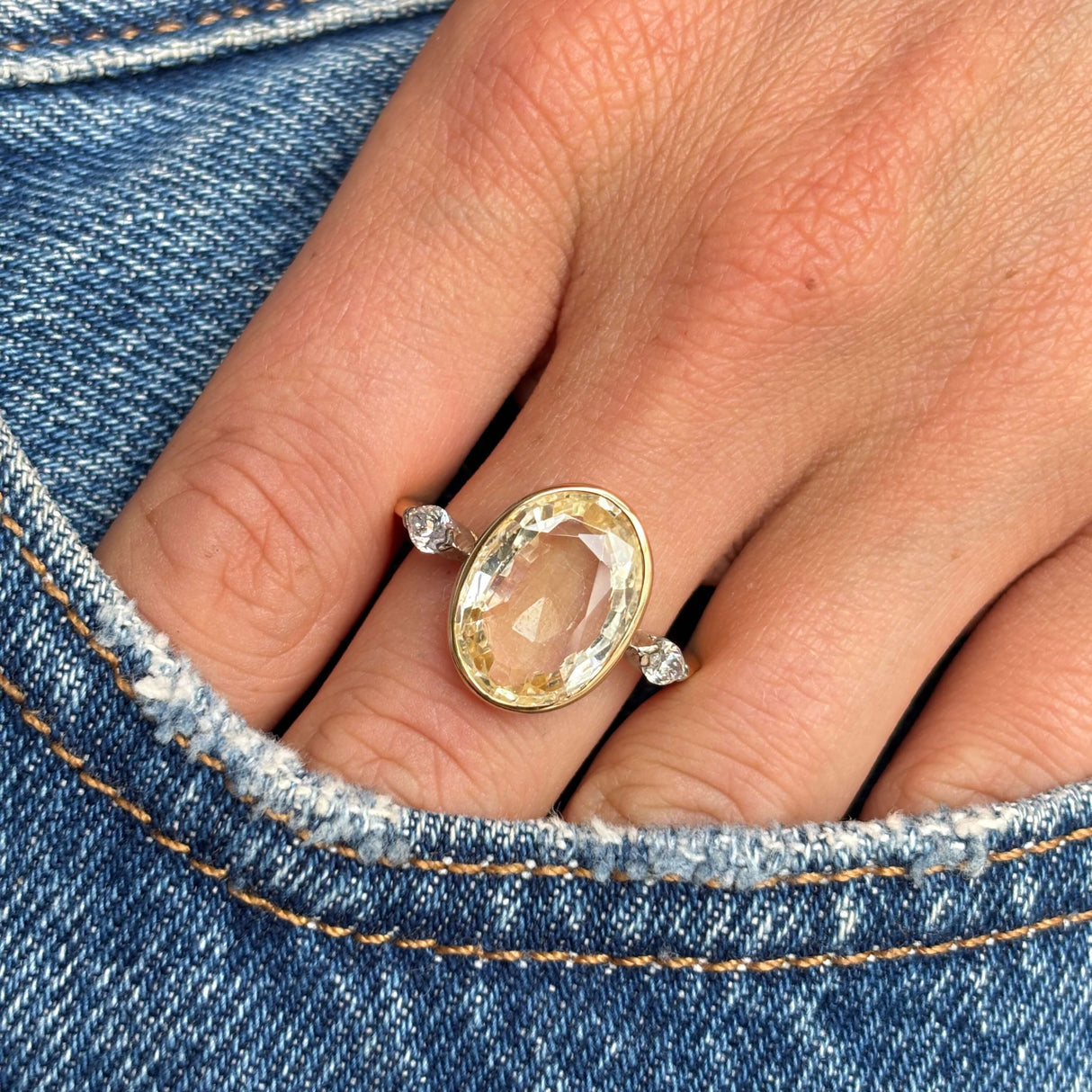 Close-up of a hand wearing a Vintage, 1980s large yellow sapphire & diamond three-stone gold ring on blue denim background