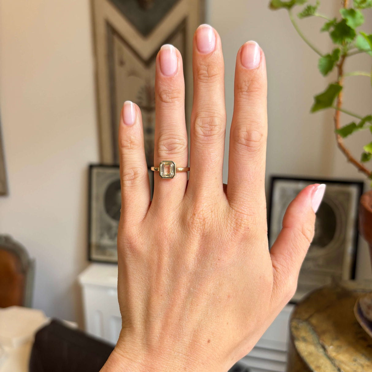 Hand wearing a gold Antique-inspired solitaire engagement ring set with a pale yellow sapphire, against a blurred indoor background.