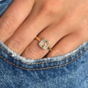 Close-up of a hand wearing a gold ring with a pale yellow sapphire, set against a denim background. Antique-inspired.