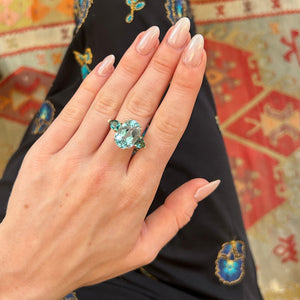 A close-up of a hand wearing a large aquamarine and green tourmaline ring,  resting on patterned black pants with blue and gold motifs, set against a warm indoor background with a patterned rug and wooden floor.