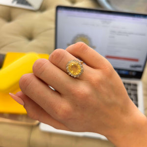 A yellow sapphire halo vintage engagement ring in yellow gold worn on a person's hand, with a laptop in the background.