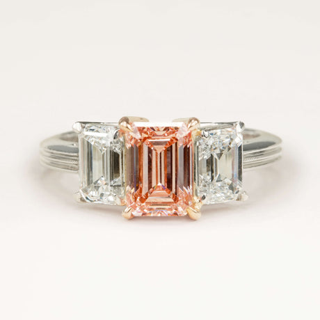 Lab-pink diamond and white diamond three-stone ring displayed on an off-white background, highlighting the pink colour contrast and refined setting.