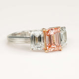 The pink diamond and white diamond three-stone ring placed on a off-white surface, details of the setting shown.