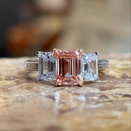 Lab-pink diamond and white diamond three-stone ring placed on a marble surface, showing the elegant proportions and gemstone arrangement.