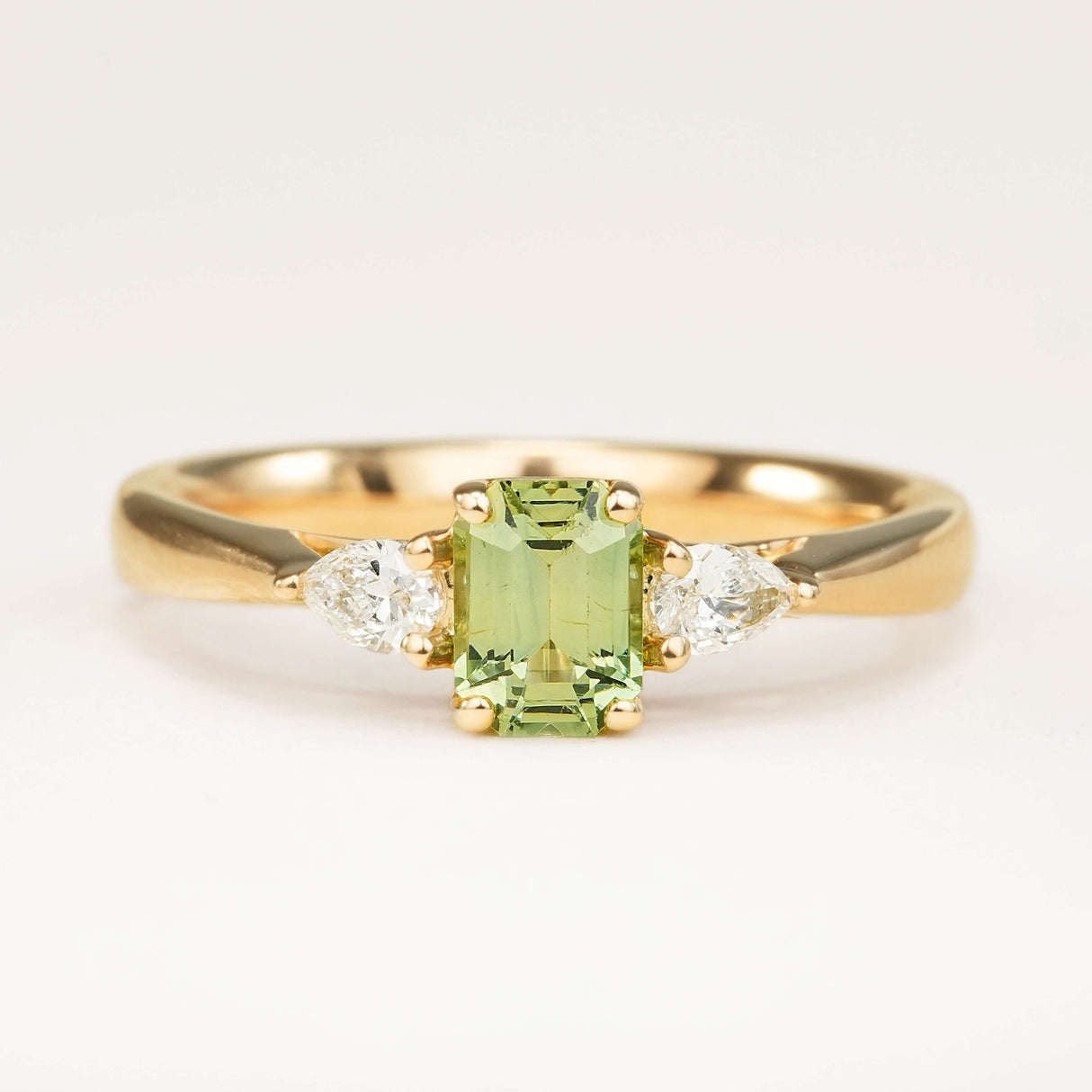 Green sapphire and diamond three-stone engagement ring set in yellow gold, displayed on an off-white background highlighting the soft green gemstone and diamond accents.