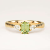 Green sapphire and diamond three-stone engagement ring set in yellow gold, displayed on an off-white background highlighting the soft green gemstone and diamond accents.