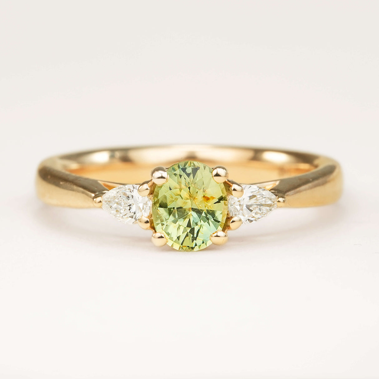 An oval-cut greeny-teal sapphire and diamond three-stone engagement ring, displayed on a clean slightly off white background.