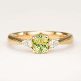 An oval-cut greeny-teal sapphire and diamond three-stone engagement ring, displayed on a clean slightly off white background.