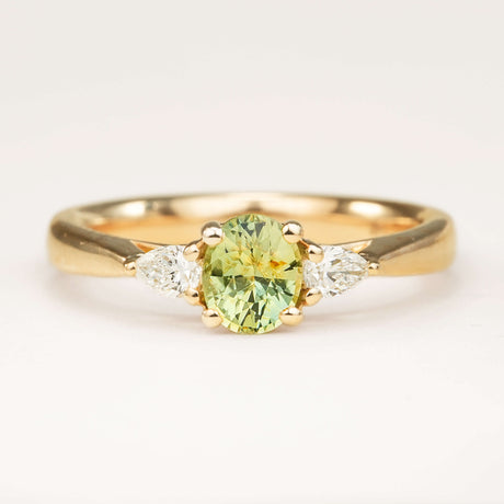 An oval-cut greeny-teal sapphire and diamond three-stone engagement ring, displayed on a clean slightly off white background.