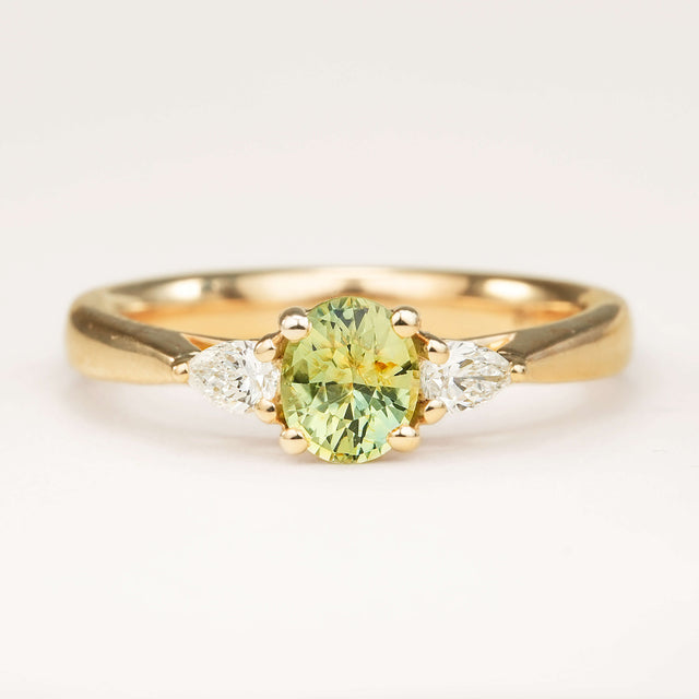 An oval-cut greeny-teal sapphire and diamond three-stone engagement ring, displayed on a clean slightly off white background.