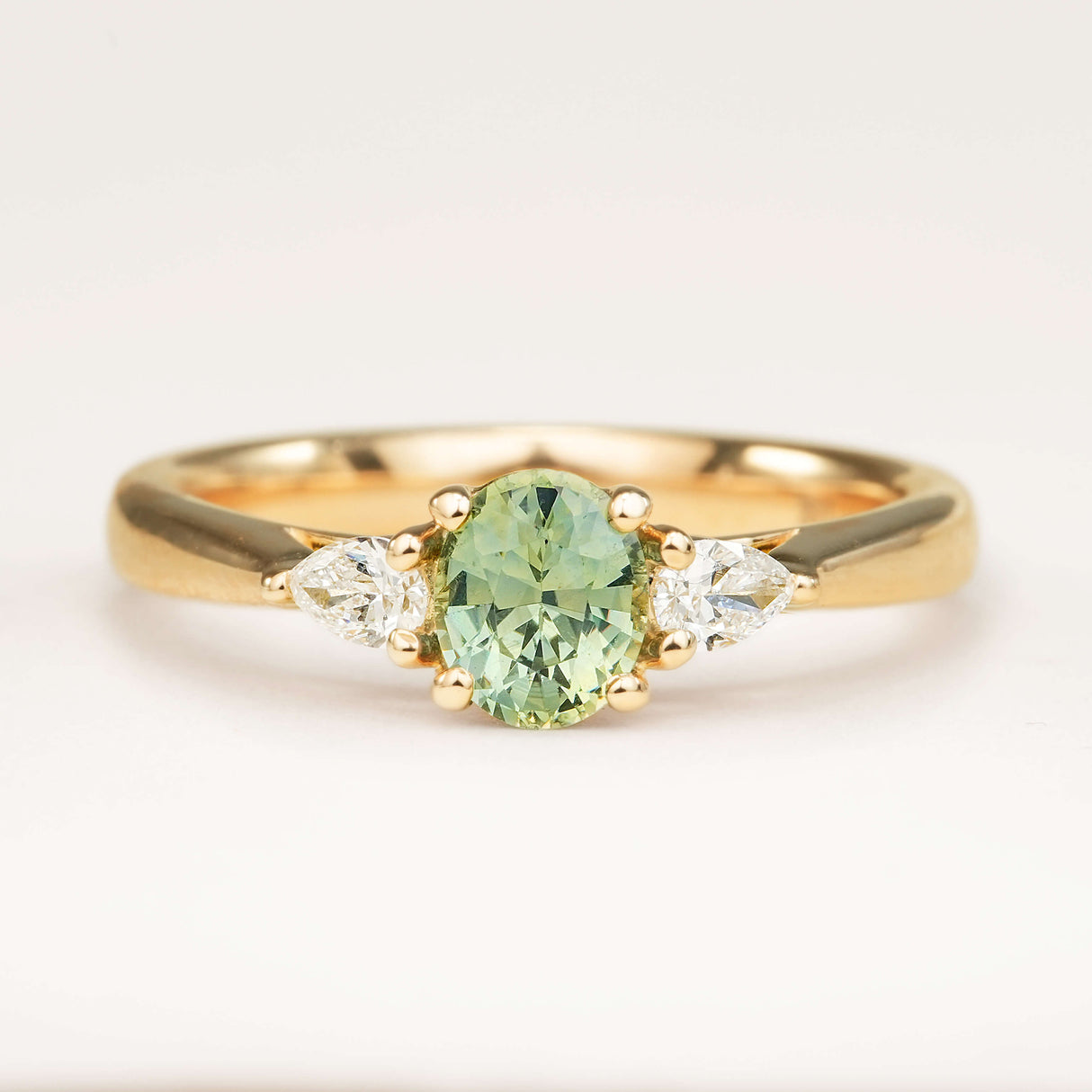 An oval-cut teal sapphire just under 1ct and pear-cut diamond three-stone engagement ring set in yellow gold, displayed on a clean white background.