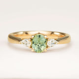 An oval-cut teal sapphire just under 1ct and pear-cut diamond three-stone engagement ring set in yellow gold, displayed on a clean white background.